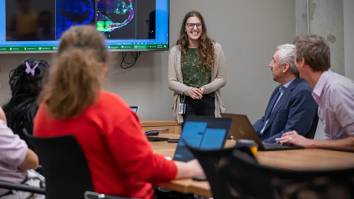 woman shares data on a tv screen to a group of individuals sitting at a board room table